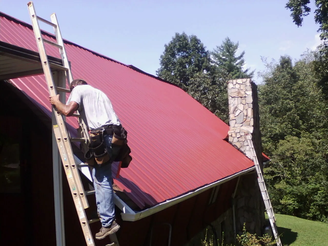 Roofer on a ladder beside a red metal roof and stone chimney — Soffit & Fascia Repair by Roofing Brothers in Rushsylvania